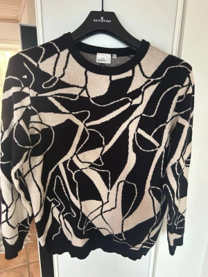 Svart och beige mönstrad stickad tröja - Säljer en svart och beige stickad tröja från Isolde med abstrakt grafiskt mönster. Tröjan har rund halsringning, långa ärmar och ribbade muddar vid ärmslut och nederkant. Perfekt för dig som gillar unika plagg med cool design.
