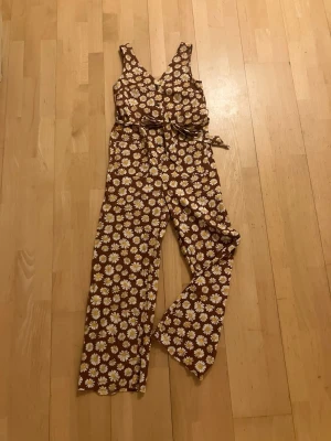 Monki Brun blommig sommardröms jumpsuit med knytband - Supersöt brun jumpsuit med vita och gula prästkragar över hela plagget. Djup v-ringning, breda axelband och knytband i midjan för snygg passform. Långa, raka ben i mjukt och lätt tyg – perfekt för dig som vill ha den här sommardrömmen! Märke Monki storlek XXS