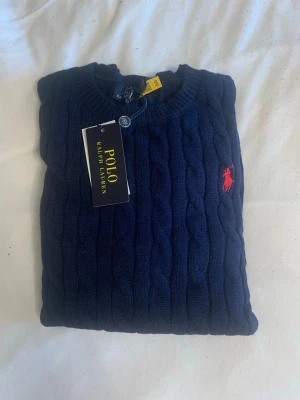 Mörkblå kabelstickad tröja Polo Ralph Lauren - Kabelstickad mörkblå tröja från Polo Ralph Lauren med rund halsringning och röd broderad logga på bröstet. Tröjan har långa ärmar och ribbade muddar vid ärmslut och nederkant. Perfekt för dig som gillar klassisk och stilren design. MÅTT: Axelbredd: 40cm. Längd: 64cm.