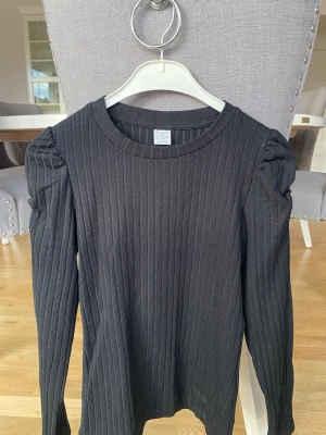 Svart ribbad tröja från Lindex - Säljer en svart långärmad ribbad tröja från Lindex med puffärmar. Tröjan har rund halsringning och är tillverkad i ett mjukt, stretchigt material. Perfekt för en stilren och enkel look. Köparen står för frakt.