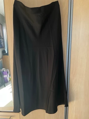 Svart långkjol i satin - Stilren svart långkjol i mjukt satinliknande material. Kjolen har en enkel, rak passform och faller snyggt längs benen. Perfekt att matcha med en crop top eller stickad tröja för en trendig look.