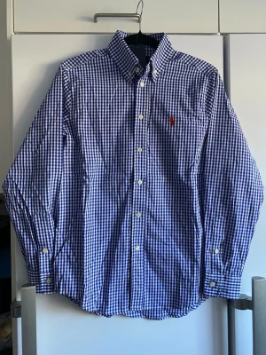 Blårutig skjorta från Polo Ralph Lauren - Klassisk blårutig skjorta från Polo Ralph Lauren med button-down krage och broderad röd logga på bröstet. Skjortan har långa ärmar och vita knappar framtill. Perfekt för dig som gillar tidlös och stilren design.