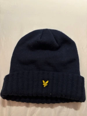 Mörkblå mössa från Lyle & Scott - Säljer en mörkblå ribbstickad mössa från Lyle & Scott med uppvikt kant och den klassiska gula örnloggan framtill. Mössan är mjuk och stretchig, perfekt för kyliga dagar. Enkel och stilren design som passar till det mesta.