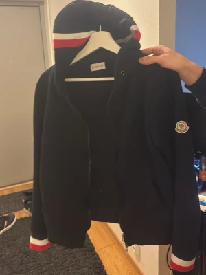 Moncler zip up - Snygg svart hoodie från Moncler med dragkedja, luvan och Moncler-logga på ärmen. Luvan har röd, vit och blå rand, och samma färger finns på muddarna. Perfekt för dig som vill ha en stilren och exklusiv look. Passar S och M. Pris går o diskuteras