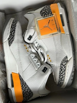 Air Jordan 3 Retro Vit/Orange - Säljer ett par Air Jordan 3 Retro sneakers i vitt läder med orange detaljer och klassiskt elephant print i grått och svart. Skorna har högt skaft, vita skosnören och ikonisk Jumpman-logga på plösen. Perfekta för dig som vill sticka ut med en unik färgkombo.