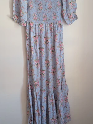 BIK Bok Blommig långklänning Storlek S. 100%Viskos - Ljusblå långklänning med rosa blommönster och smockad överdel. Klänningen har puffiga trekvartsärmar och volang nertill för en romantisk look. Tillverkad i ett lätt och luftigt material, perfekt för varma dagar.