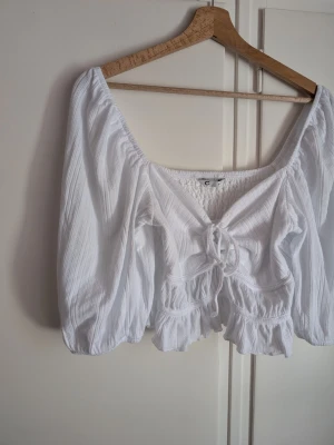 Cubus Vit croppad topp med snörning Storlek M - Söt vit croppad topp med långa puffärmar och snörning framtill. Toppen har volangdetaljer vid midjan och ärmarna, samt en lätt ribbad struktur i tyget. Perfekt för en trendig och luftig look.