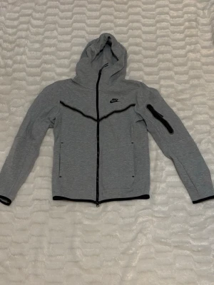 Grå Nike tech  - Snygg grå hoodie från Nike med hel dragkedja och svarta detaljer. På ena armen finns defekter 