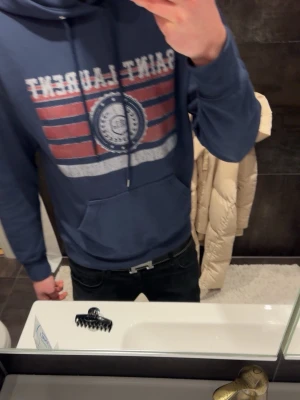 Blå hoodie från Saint Laurent - Säljer en blå hoodie från Saint Laurent med tryckt logga och ränder i rött och vitt på bröstet. Hoodien har huva med dragsko och en stor magficka framtill. Perfekt för dig som gillar streetwear och vill ha något bekvämt och snyggt.