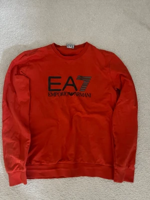Röd sweatshirt från Emporio Armani - Säljer en röd sweatshirt från Emporio Armani EA7 med svart logga på bröstet. Tröjan har rund halsringning, långa ärmar och ribbade muddar vid ärmslut och nederkant. Perfekt för en avslappnad och trendig look.