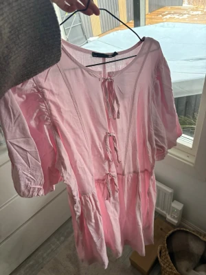 Ljusrosa kort klänning med knyt - Söt ljusrosa kort klänning med puffärmar och tre dekorativa knyt framtill. Tillverkad i mjuk bomull som känns skön mot huden. Klänningen har en avslappnad passform och lätt rynkad kjol för en trendig look.