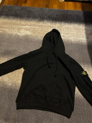 Svart hoodie från Stone Island - Storlek M eller mindre L. Säljer en svart hoodie från Stone Island med klassisk patch på vänster ärm och stor magficka framtill. Perfekt för dig som gillar streetwear och vill ha en stilren look.