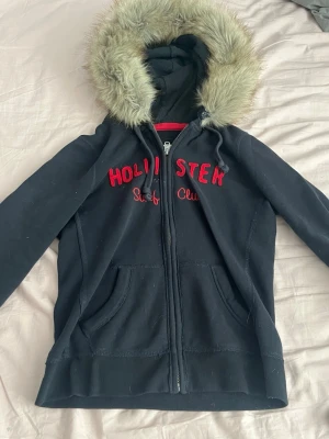 Svart hoodie Hollister med fuskpäls - Mörk blå hoodie från Hollister med dragkedja, röda broderade bokstäver och texten 'Surf Club' på bröstet. Stor huva med fuskpäls runt kanten och två fickor framtill. Perfekt för en chill och avslappnad stil.