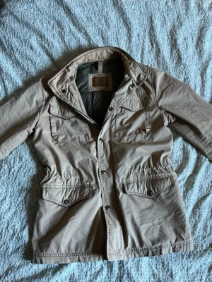 Beige vindjacka med fickor - Stilren beige vindjacka med fyra stora fickor framtill och knappstängning. Jackan har hög krage och justerbar midja för en snygg passform. Tillverkad i lätt och slitstarkt material, perfekt för blåsiga dagar.