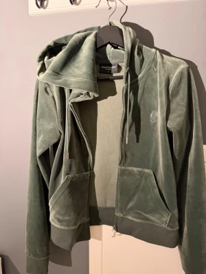 Grön velour loungewear-set Juicy Couture - Mysigt loungewear-set från Juicy Couture i mjuk grön velour. Setet består av en zip-hoodie med fickor och dragsko samt matchande byxor med resårmidja och fickor. Perfekt för chill dagar hemma eller när du vill vara extra bekväm.