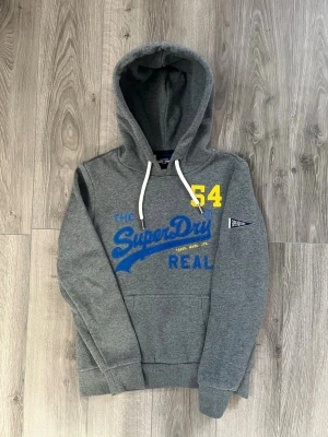 Grå Superdry hoodie med tryck - Snygg grå hoodie från Superdry med blått och gult tryck framtill och vita dragsnören i huvan🤩 passar nån i storlek Xs och inga fel😇 hör av er vid snabbaffär samt funderingar 