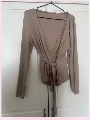 Beige omlottopp med knytband - Trendig beige omlottopp med långa ärmar och knytband i midjan. Toppen har en djup v-ringning och är tillverkad i ett mjukt, tunt material som känns skönt mot huden. Perfekt att styla med jeans eller kjol för en snygg look.💞LÄGG GÄRNA PRISFÖRSLAG !