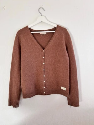 Brun kofta från Odd Molly - Mysig brun finstickad kofta från Odd Molly med v-ringning och pärlknappar framtill. Koftan har ribbade muddar, dragkedjor vid ärmsluten och en liten patch med logga nedtill. Perfekt för lager på lager och en avslappnad stil.