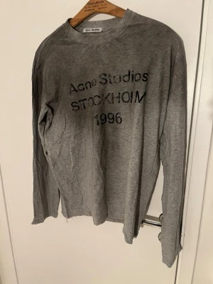 Grå långärmad topp Acne Studios - Snygg grå långärmad topp från Acne Studios med svart texttryck på bröstet där det står 'Acne Studios STOCKHOLM 1996'. Tillverkad i mjukt material med avslappnad passform och rund halsringning. Perfekt för en stilren och enkel look.