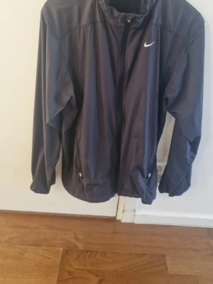 Svart vindjacka från Nike - Snygg svart vindjacka från Nike med dragkedja framtill och två fickor med dragkedja. Två fickor på insidan av jackan. Jackan har en minimalistisk design med Nike-logga på bröstet och hög krage. Tillverkad i lätt och vindtätt material, perfekt för blåsiga dagar.(Pris går att ändra