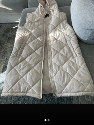 Ljusbeige lång väst med huva - Säljer en ljusbeige lång Täckväst med huva och dragkedja framtill. Västen har ett quiltat mönster i stora rutor och praktiska fickor med dragkedja. Perfekt för lager på lager och ger en clean look till din outfit.