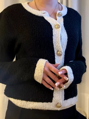 Svart och vit bouclékofta med guldknappar - Stilren svart bouclékofta med vita kontrastkanter och dekorativa guldknappar framtill. Koftan har långa ärmar, rund halsringning och vita detaljer vid ärmslut, nederkant och fickor. Perfekt för en chic och trendig look.