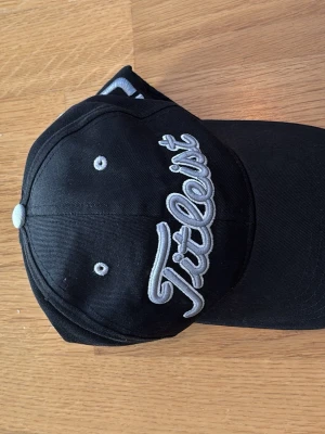 Svart Titleist keps med brodyr - Säljer en svart keps från Titleist med stor broderad logga i vitt och grått framtill. Kepsen har klassisk böjd skärm och ventilationshål med kontrastfärg. Perfekt för dig som gillar sportig stil och vill ha en snygg accessoar.