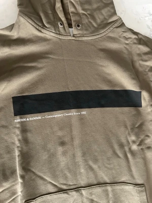 Grön hoodie från Samsøe & Samsøe - Snygg grön hoodie från Samsøe & Samsøe med svart rektangulärt tryck och texten 'Contemporary Classics Since 1993' på bröstet. Klassisk känguruficka framtill och huva. Perfekt för en chill och stilren look.