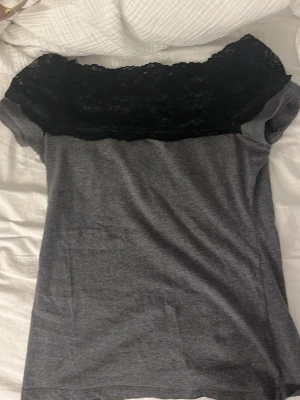 Grå topp med svart spetsdetalj - Snygg grå topp med svart spets upptill som ger en elegant touch. Toppen har korta ärmar och en mjuk, bekväm passform. Perfekt för dig som gillar stilrena plagg med lite extra detalj.