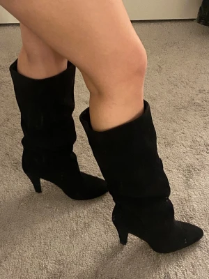 Svarta mocka boots - Säljer ett par svarta höga stövlar i mockaimitation. Stövlarna har smal klack som ger en snygg siluett och passar perfekt till kjol eller klänning. Aldrig använda!