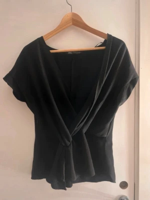 Zara blus - Snygg svart omlottopp med knyt i midjan och v-ringad hals. Toppen har korta ärmar och en smickrande passform som framhäver midjan. Perfekt till jeans eller kjol för en trendig look.