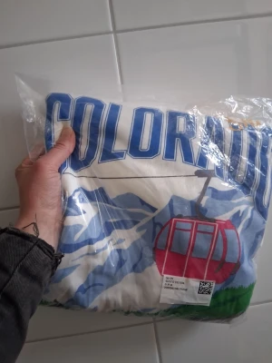 Colorado hoodie med berg och linbana - Säljer en vit hoodie med stor blå text 'COLORADO' över bröstet och ett färgglatt tryck av berg och en röd linbana. Hoodien har huva och en avslappnad passform, perfekt för dig som gillar streetwear och coola tryck.