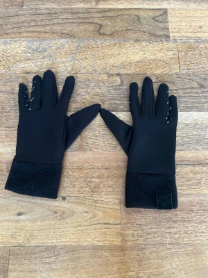 Svarta fleecefodrade ridhandskar - Svarta handskar med mjukt fleecefoder och greppmönster i vitt på insidan av fingrar och handflata. Perfekta för kyliga dagar och har en enkel, stilren design. Ger bra grepp åt tyglarna.
