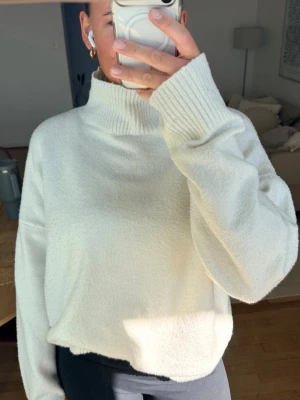 Vit stickad polotröja - Mysig vit stickad polotröja med oversized passform och breda ribbade muddar vid ärmslut och hals. Tröjan har en hög krage och är gjord i ett mjukt material som känns skönt mot huden. Perfekt för kalla dagar och enkel att styla till olika outfits.