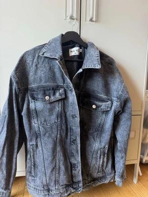 Svartgrå jeansjacka från NA-KD - Snygg svartgrå jeansjacka från NA-KD med klassisk rak passform. Jackan har två bröstfickor med knappar, framfickor och markerad krage. Använts en del men i bra skick.
