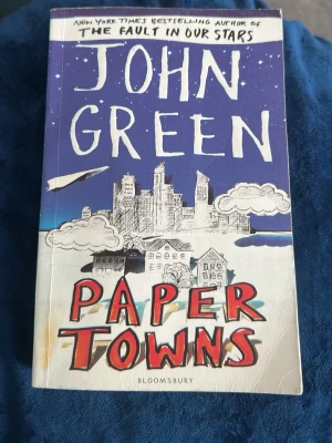 Paper Towns av John Green - En ungdomsroman på engelska av John Green med färgstarkt omslag i blått, vitt och rött. Boken har ett tecknat stadslandskap och pappersflygplan på framsidan. Perfekt för dig som gillar mysterier, kärlek och roadtrips i amerikansk high school-miljö.