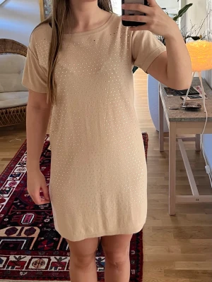 Kortklänning med stenar - Snygg beige rosa kortklänning med korta ärmar och rund halsringning. Klänningen har små pärlor framtill som ger ett elegant intryck. Tillverkad i mjukt material som känns skönt mot huden. Perfekt för dig som gillar en enkel men ändå stilren look.