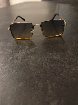 Svart-guldiga Louis Vuitton glasögon - Snygga solglasögon med fyrkantiga glas och guldfärgade metallbågar. Glasen är mörka med en lätt toning och skalmarna har en kedjedetalj. Passar perfekt för dig som vill ha en trendig accessoar med lyxig känsla.