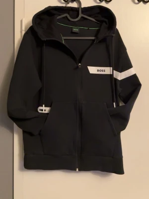 Svart hoodie med dragkedja från BOSS - Svart hoodie från BOSS med vit logga på bröstet och vit rand på ena ärmen. Modellen har dragkedja framtill, två fickor och justerbar huva med snören. Tillverkad i mjukt material som känns skönt mot huden. Perfekt för en avslappnad och stilren look.