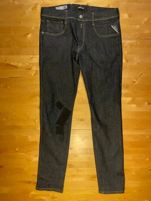 Mörkblå jeans från Replay - Snygga mörkblå jeans från Replay med raka ben och klassisk femficksmodell. Jeansen har kontrastsömmar och patchdetaljer på vänster ben. Tillverkade i slitstark denim med läderpatch bak i midjan. Perfekta för en trendig och avslappnad stil.