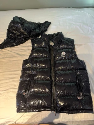 Svart glansig dunväst från Moncler - Snygg svart dunväst från Moncler med glansigt material och avtagbar huva. Västen har två dragkedjefickor framtill och Moncler-logga på bröstet. Perfekt för lager på lager och streetwear-vibe.