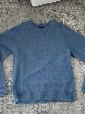 Blå stickad tröja från Uniqlo - Mysig blå stickad tröja från Uniqlo med rund halsringning och ribbade muddar vid ärmslut och nederkant. Enkel och stilren design. Materialet känns mjukt och behagligt mot huden.