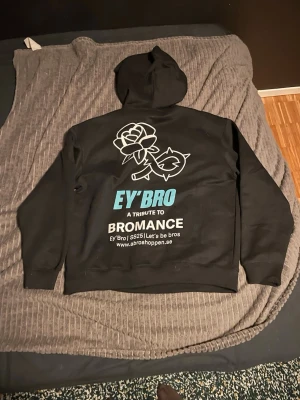 Svart hoodie från EY'BRO med tryck - Svart hoodie från EY'BRO med stor vit och blå text på ryggen: 'A tribute to bromance' samt en ros och hjärta-illustration. Klassisk känguruficka framtill och liten logga på bröstet. Mjuk och skön, perfekt för chill dagar. Helt ny!
