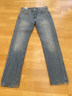 Levi's 502 raka blå jeans - Klassiska blå jeans från Levi's med rak passform och fem fickor. Jeansen har en ljus tvätt med slitningar på låren och knäna, samt den ikoniska röda Levi's-lappen på bakfickan. Tillverkade i slitstark denim, perfekta för en avslappnad stil.
