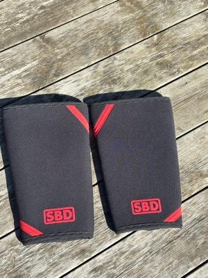 SBD knäskydd svart/röd - Säljer ett par svarta SBD knäskydd med röda detaljer. Perfekta för styrketräning och gym, ger stabilitet och stöd vid tunga lyft. Tillverkade i slitstarkt syntetmaterial och har en clean design med SBD-logga framtill. Använda uppskattningsvis 4-5 gånger och har inga täcken på användning