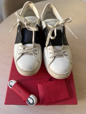 Valentino sneakers  - Snygga sneakers från Valentino i vitt läder med svart över mitten och nitar på hälen. Klassisk låg modell med vita snören och gummisula. Kommer med extra original skosnören.