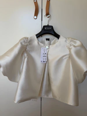 Snygg fest topp - Snygg överdel i glansigt satinmaterial i offwhite. Jackan har vida, korta ärmar och en enkel knäppning vid halsen. Perfekt för att lyfta en outfit med en elegant touch. Rak passform och rundad halsringning.