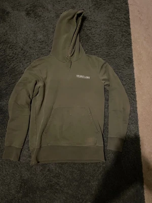 Helmut Lang hoodie  - Snygg olivgrön hoodie från Helmut Lang med vit logga tryckt på bröstet. Klassisk känguruficka framtill och ribbade muddar vid ärmar och nederkant. Tillverkad i mjukt bomullsmaterial och har en stor huva för extra komfort.