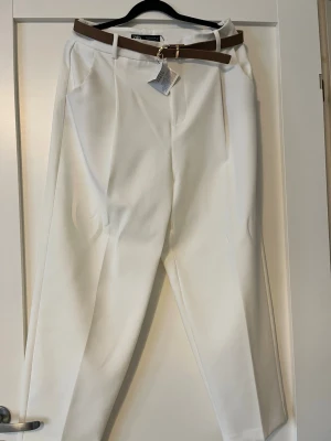Vita kostymbyxor från ZARA - Snygga vita kostymbyxor från ZARA med hög midja och raka ben. Byxorna har pressveck, sidofickor och medföljer ett brunt bälte i midjan. Perfekta för en clean och stilren look. Materialet känns mjukt och har en lätt struktur. Ankellånga. Köpta i Spanien