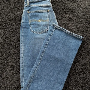 Blå utsvängda jeans från Nudie Jeans co W26 L30 - Säljer ett par klassiska blå jeans från Nudie Jeans med rak passform och femficksmodell. Jeansen är tillverkade i ekologisk eller återvunnen bomull och har snygga kontrastsömmar samt en diskret logga på bakfickan. Perfekta för en avslappnad och stilren look.